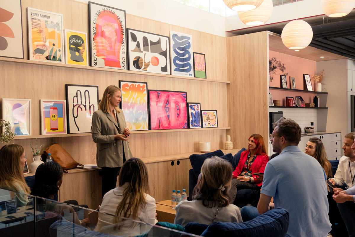 A woman stands talking to a group seated in a modern room. The room features a wall adorned with colorful abstract and graphic art pieces. The space has a cozy and inviting atmosphere with blue couches and soft lighting.